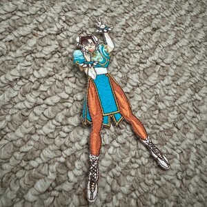 Może przedstawiać: Haftowana naszywka przedstawiająca postać w niebiesko-pomarańczowym stroju z białymi akcentami. Naszywka przedstawia kobietę w dynamicznej pozie z uniesionymi ramionami. Naszywka znajduje się na teksturowanej, neutralnej powierzchni.