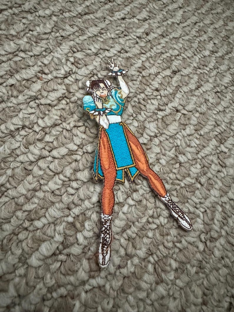 Może przedstawiać: Haftowana naszywka przedstawiająca postać w niebiesko-pomarańczowym stroju z białymi akcentami. Naszywka przedstawia kobietę w dynamicznej pozie z uniesionymi ramionami. Naszywka znajduje się na teksturowanej, neutralnej powierzchni.
