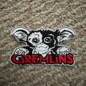 May include: Embroidered patch featuring a Gremlin character with large ears, a mischievous grin, and large eyes. The patch has a black and white design with the word "GREMLINS" in red lettering.