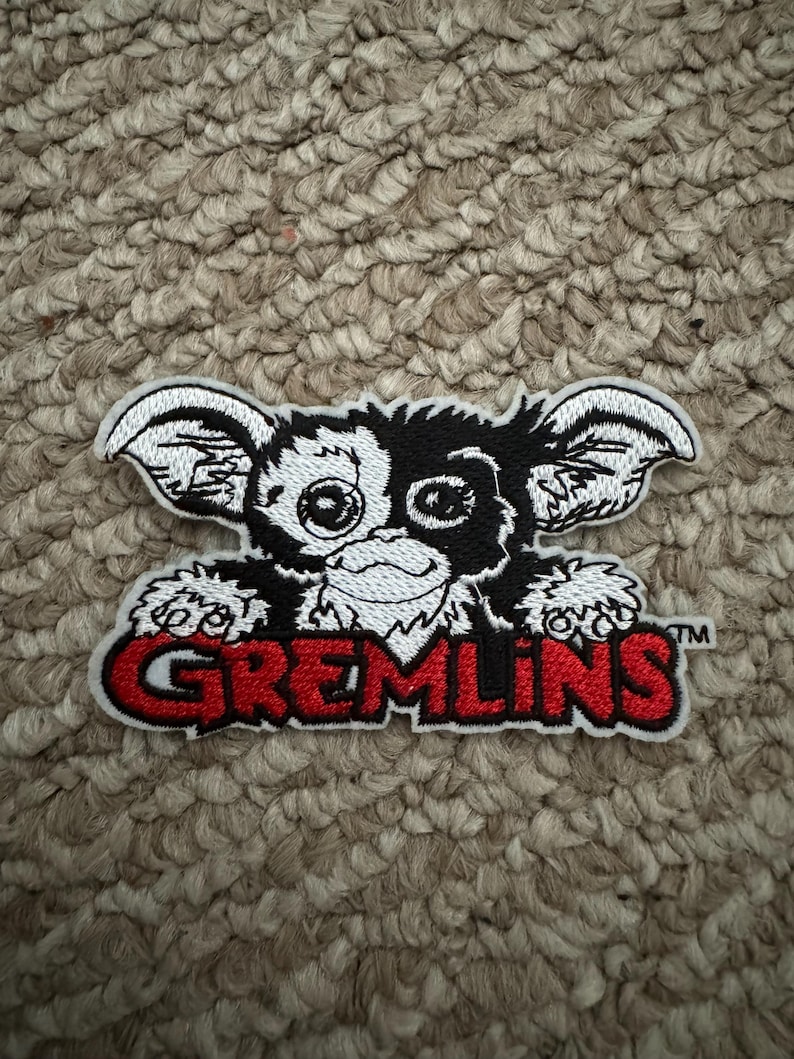 May include: Embroidered patch featuring a Gremlin character with large ears, a mischievous grin, and large eyes. The patch has a black and white design with the word "GREMLINS" in red lettering.
