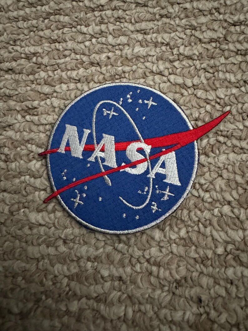 Peut inclure: Un &eacute;cusson NASA bleu circulaire avec du texte brod&eacute; blanc et un trait rouge. L'&eacute;cusson pr&eacute;sente le logo de la NASA avec des &eacute;toiles et un design en ligne incurv&eacute;e. Cet &eacute;cusson est probablement destin&eacute; aux v&ecirc;tements ou aux accessoires.