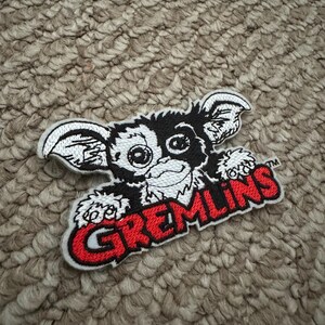 May include: Embroidered patch of a Gremlin character with a black and white face, large ears, and a grin. The word "GREMLINS" is in red letters with a white outline. The patch is on a textured, neutral-toned surface.