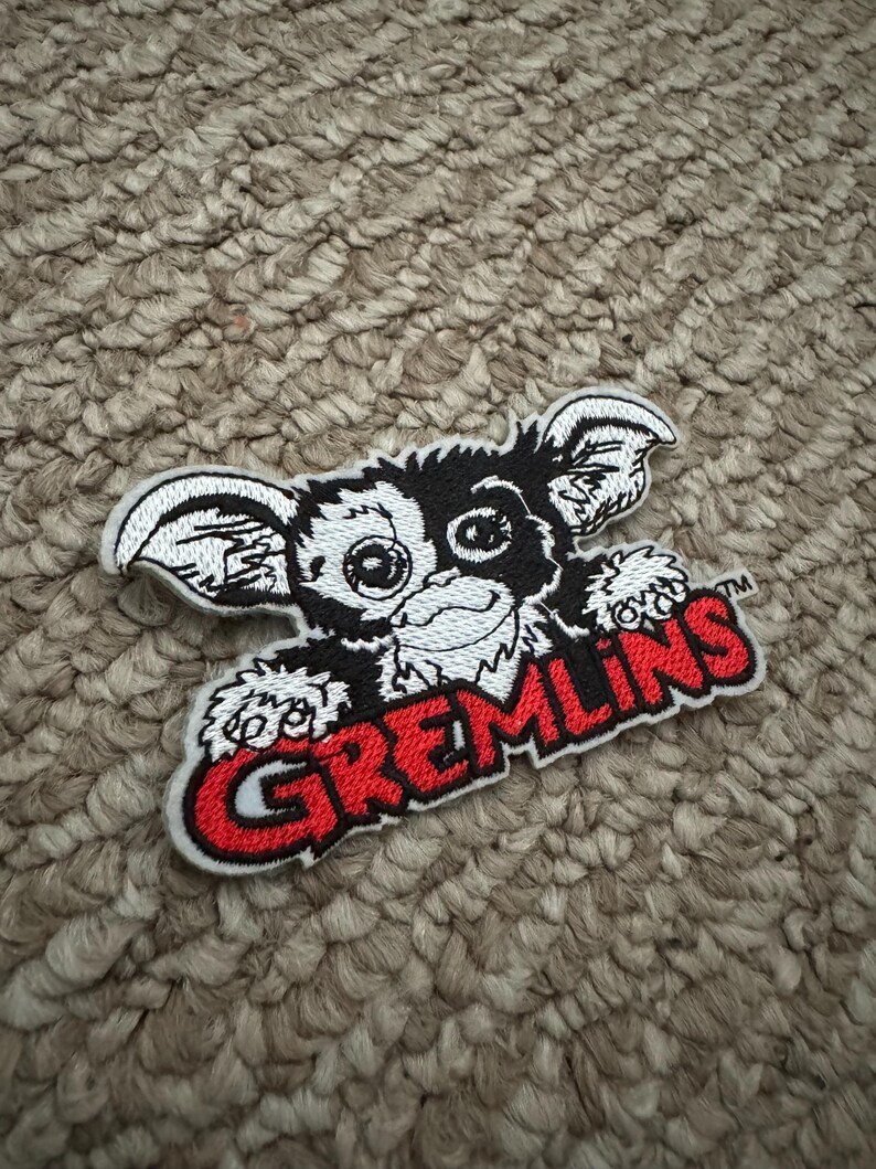 May include: Embroidered patch of a Gremlin character with a black and white face, large ears, and a grin. The word "GREMLINS" is in red letters with a white outline. The patch is on a textured, neutral-toned surface.