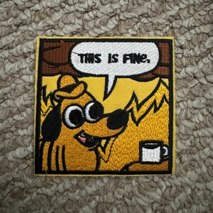 May include: Embroidered patch showing a cartoon dog with a hat, holding a coffee cup, and the text "THIS IS FINE." The dog is yellow with black accents, set against a yellow and brown background. The patch is square with a yellow border.
