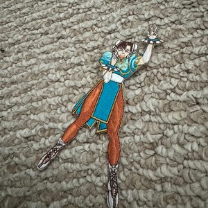 Może przedstawiać: Haftowana naszywka przedstawiająca postać w dynamicznej pozie. Postać nosi niebiesko-pomarańczowy str&oacute;j z białymi detalami. Naszywka jest bogato zdobiona, prezentując motyw gry walki.