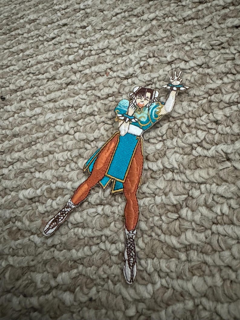Może przedstawiać: Haftowana naszywka przedstawiająca postać w dynamicznej pozie. Postać nosi niebiesko-pomarańczowy str&oacute;j z białymi detalami. Naszywka jest bogato zdobiona, prezentując motyw gry walki.