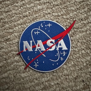 May include: A circular patch featuring the NASA logo. The patch has a blue background with the word "NASA" in white, a red swoosh, and white stars and curved lines. The patch is embroidered.