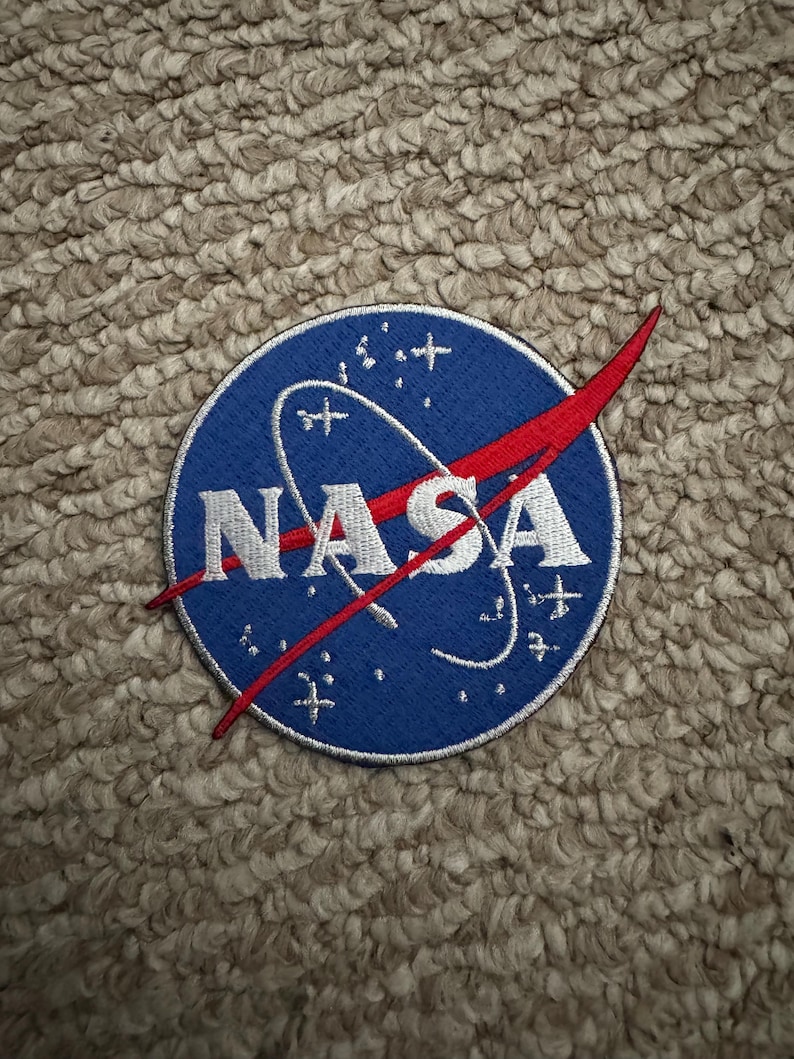 Peut inclure: Un &eacute;cusson circulaire avec le logo de la NASA. Le patch a un fond bleu avec le mot "NASA" en blanc, un trait rouge et des &eacute;toiles et lignes blanches. L'&eacute;cusson est brod&eacute;.