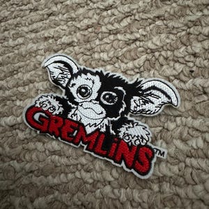May include: Embroidered patch of a Gremlin character in black and white, with large ears and eyes. The word "GREMLINS" is in red across the bottom. The patch is on a textured, neutral-toned surface.