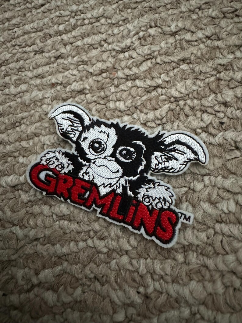 May include: Embroidered patch of a Gremlin character in black and white, with large ears and eyes. The word "GREMLINS" is in red across the bottom. The patch is on a textured, neutral-toned surface.