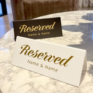 May include: Two white and black table cards with gold lettering that say "Reserved" and "Name & Name". The cards are on a white marble table.