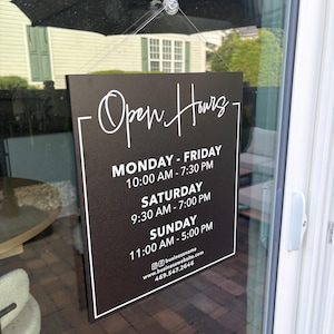 May include: A rectangular black sign with white text hanging on a glass door. The sign reads Open Hours, Monday-Friday 10:00 AM-7:30 PM, Saturday 9:30 AM-7:00 PM, Sunday 11:00 AM-5:00 PM, along with a social media handle, website, and phone number 469.547.2646.