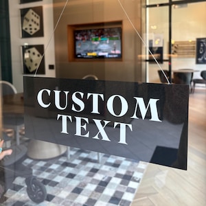 May include: A black rectangular sign with the words "CUSTOM TEXT" in large white capital letters. The sign is hanging by thin wires, and the background shows a room with a television and a checkered rug.