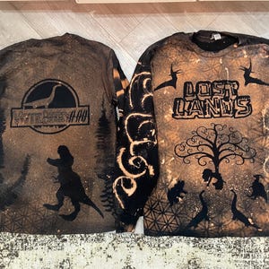 Puede incluir: Dos camisas de manga larga con diseños de decoloración con lejía. La camisa de la izquierda presenta una silueta negra de un dinosaurio con el texto "Vote Berry 4110". La camisa de la derecha presenta un árbol con el texto "Lost Lands".
