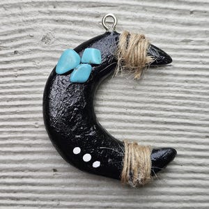 May include: Black crescent moon pendant with turquoise-colored stones and white dots. The pendant is adorned with twine and a silver-colored loop for hanging. The surface has a textured, glossy finish.