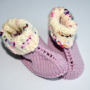 May include: A pair of hand-knitted booties featuring a solid light purple base with a textured knit pattern. The cuffs are made from a thick, cream-colored yarn speckled with vibrant multi-colored flecks, creating a distinct contrast.