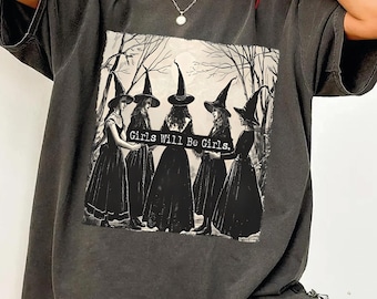 Può includere: Una t-shirt a maniche corte color pepe con una grafica in bianco e nero di cinque figure che indossano cappelli da strega e abiti lunghi in cerchio. Il testo "Girls Will Be Girls." è stampato al centro del disegno grafico.