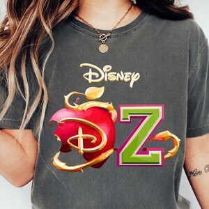 Puede incluir: Camiseta gris con el logo de Disney, un diseño de manzana roja y la letra "Z" en verde y rosa. La palabra "pepper" está impresa en la parte inferior de la camiseta.