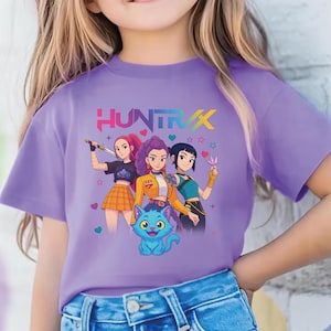 May include: A young person wearing a purple t-shirt with the word "HUNTRAX" in colorful letters. The shirt features a graphic of three animated figures and a blue cat. The person is also wearing sunglasses.