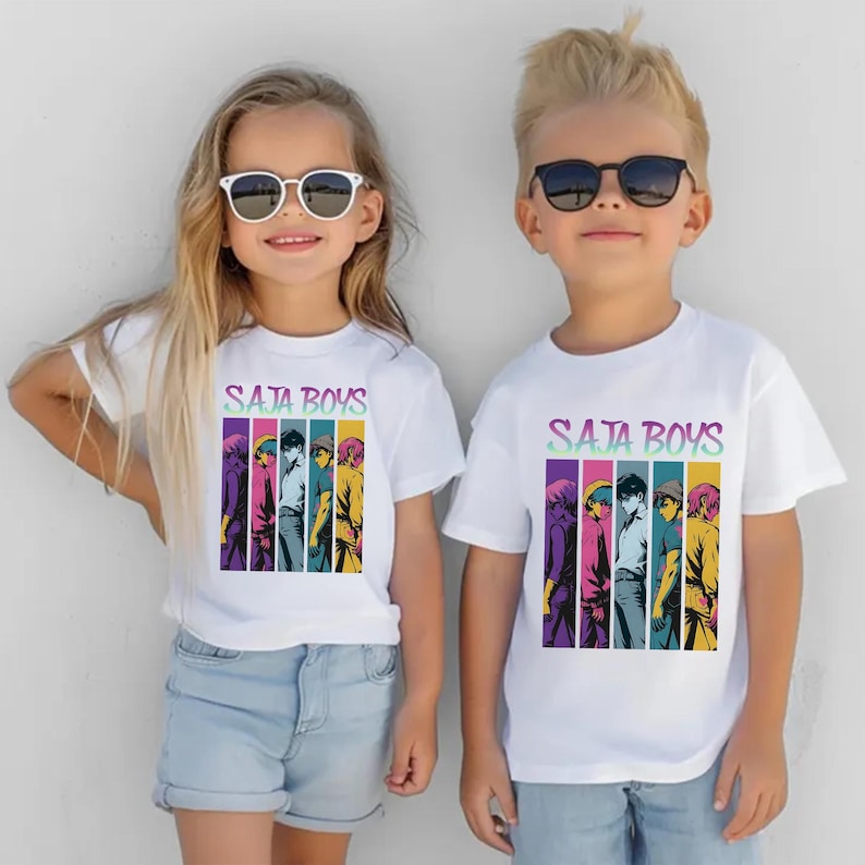 May include: Two children wearing white t-shirts with the text "Saja Boys" in a colorful, gradient font. Below the text is a vertical graphic of six anime-style figures. Both children are wearing sunglasses.