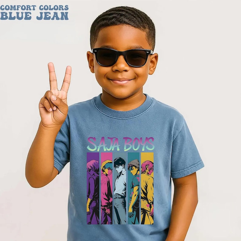 May include: A blue jean colored t-shirt with the text "SAJA BOYS" and a graphic of five stylized figures. The shirt also has the text "COMFORT COLORS" and "BLUE JEAN" at the top. The person in the image is wearing black sunglasses and making a peace sign.