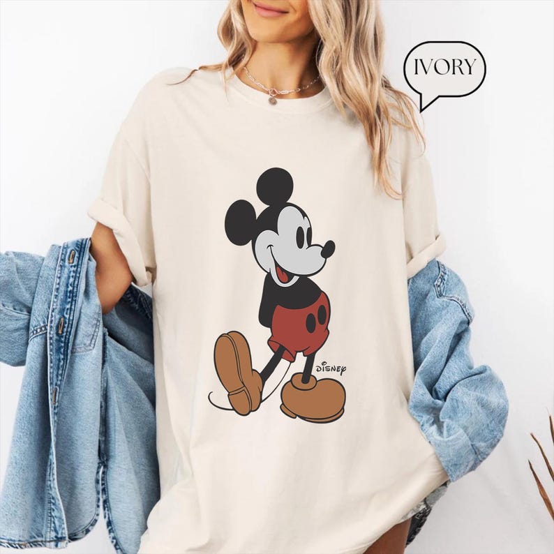 Op de afbeelding: Een wit T-shirt met een zwart-witte afbeelding van Mickey Mouse die een rode broek draagt. De tekst "Disney" is onder de afbeelding gedrukt. Het T-shirt is gemarkeerd met "IVORY".
