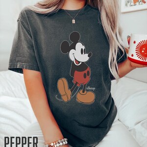 Op de afbeelding: Een donkergrijs T-shirt met een vintage afbeelding van Mickey Mouse in een rode broek en gele schoenen. Het shirt heeft het woord "Disney" in wit onder de afbeelding.