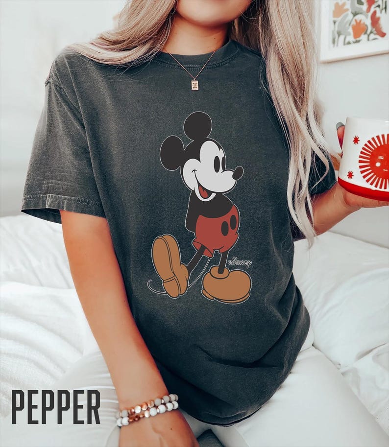Op de afbeelding: Een donkergrijs T-shirt met een vintage afbeelding van Mickey Mouse in een rode broek en gele schoenen. Het shirt heeft het woord "Disney" in wit onder de afbeelding.