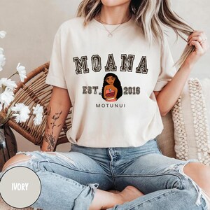 May include: A white t-shirt with a black graphic print of the Disney character Moana. The text "Moana Est. 2016 Motunui" is printed on the shirt.