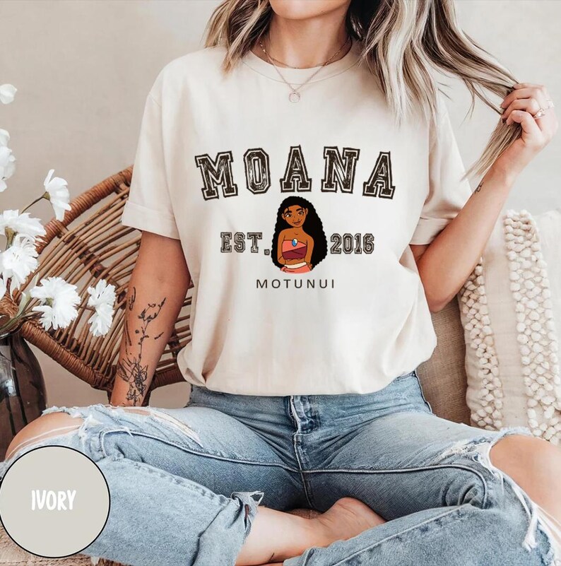 May include: A white t-shirt with a black graphic print of the Disney character Moana. The text "Moana Est. 2016 Motunui" is printed on the shirt.