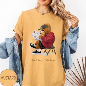 May include: A mustard yellow t-shirt with a graphic of a cartoon bear wearing a baseball cap and reading a newspaper. The text "Read More, Talk Less" is printed below the bear.