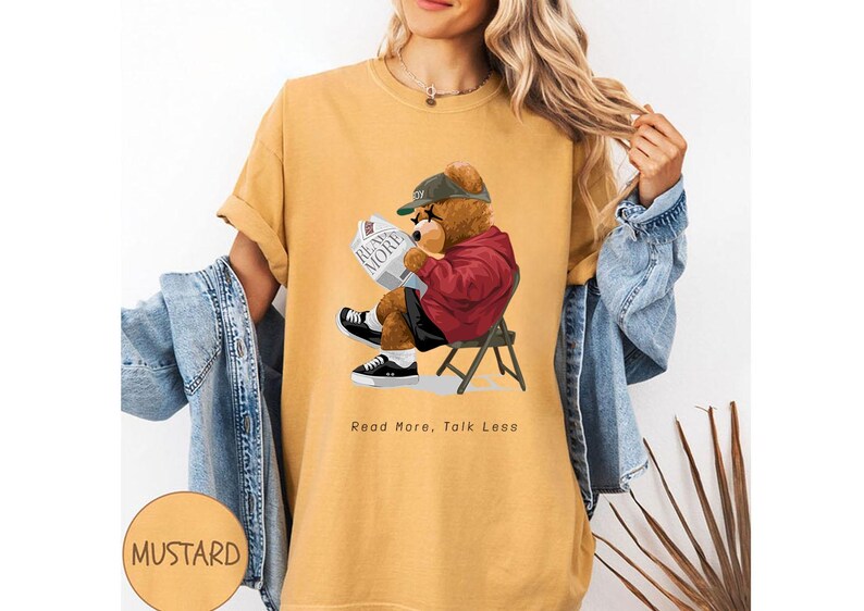 May include: A mustard yellow t-shirt with a graphic of a cartoon bear wearing a baseball cap and reading a newspaper. The text "Read More, Talk Less" is printed below the bear.