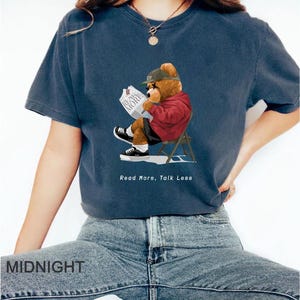 May include: A navy blue t-shirt with a cartoon illustration of a bear wearing a red jacket and a baseball cap, sitting in a chair and reading a newspaper. The text "Read More, Talk Less" is printed below the bear.