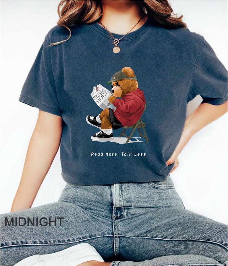 May include: A navy blue t-shirt with a cartoon illustration of a bear wearing a red jacket and a baseball cap, sitting in a chair and reading a newspaper. The text "Read More, Talk Less" is printed below the bear.