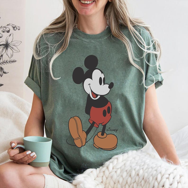 Op de afbeelding: Een groen T-shirt met een vintage Mickey Mouse-afbeelding. De afbeelding toont Mickey Mouse in een rood-zwarte outfit, lachend en lopend. De tekst "Disney" is onder de afbeelding gedrukt.