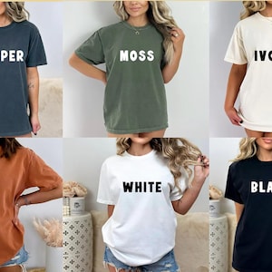 May include: Six different colored t-shirts with various text printed on the front. The colors are black, white, ivory, moss green, pepper grey, and yam orange. The text on the shirts reads "BLACK", "WHITE", "IVORY", "MOSS", "PEPPER", and "YAM".