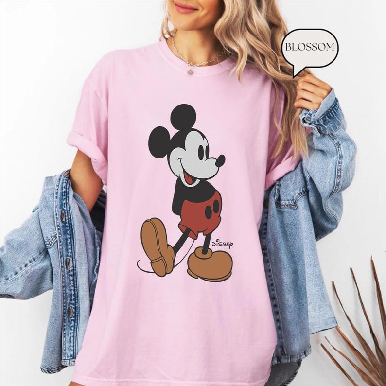 Op de afbeelding: Roze T-shirt met een afbeelding van Mickey Mouse in een klassieke pose. De afbeelding is zwart en rood met het woord "Disney" eronder.