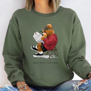 May include: Green sweatshirt with a cartoon illustration of a teddy bear wearing a baseball cap and sunglasses, reading a newspaper with the text "Read More" on it. The bear is sitting on a chair and the text "Read More, Talk Less" is printed below the image.