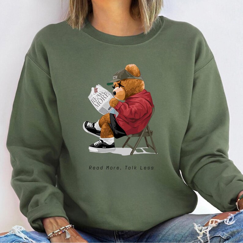 May include: Green sweatshirt with a cartoon illustration of a teddy bear wearing a baseball cap and sunglasses, reading a newspaper with the text "Read More" on it. The bear is sitting on a chair and the text "Read More, Talk Less" is printed below the image.