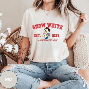 May include: Ivory t-shirt with a graphic print of Snow White from the Disney movie Snow White and the Seven Dwarfs. The graphic includes the text "Snow White", "Est. 1937", and "A Kingdom Far Away".