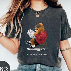 May include: A dark gray t-shirt with a cartoon illustration of a bear wearing a red jacket and a green baseball cap. The bear is sitting on a chair and reading a newspaper that says "Read More". The text "Read More, Talk Less" is printed below the bear.