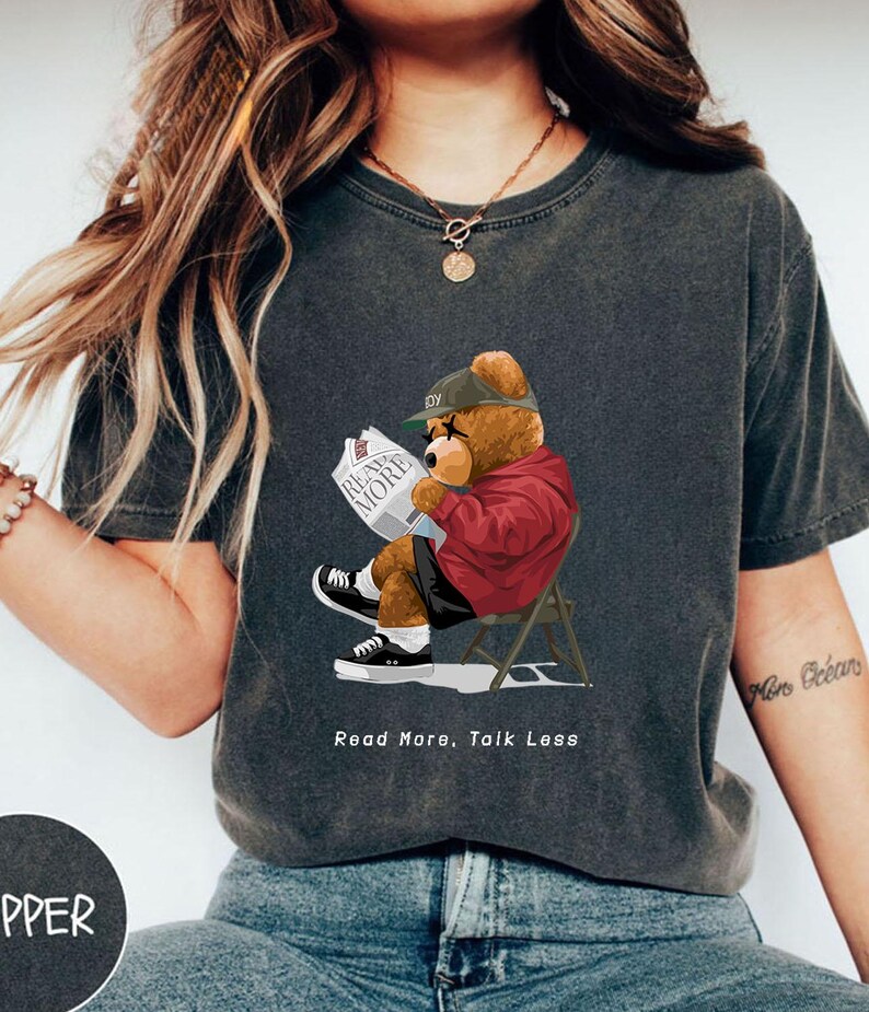 May include: A dark gray t-shirt with a cartoon illustration of a bear wearing a red jacket and a green baseball cap. The bear is sitting on a chair and reading a newspaper that says "Read More". The text "Read More, Talk Less" is printed below the bear.