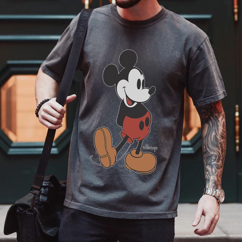 Op de afbeelding: Een antracietgrijs T-shirt met een vintage-illustratie van Mickey Mouse in zwart-wit. De illustratie toont Mickey Mouse terwijl hij loopt met een glimlach, met een rode broek en zwarte schoenen. De tekst "Disney" is in wit onder Mickey Mouse gedrukt.