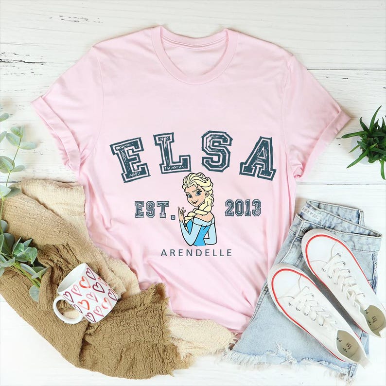 May include: Pink t-shirt with a blue and white graphic of Elsa from Frozen. The graphic includes the text "Elsa Est. 2013 Arendelle".