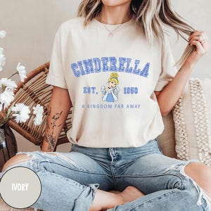 May include: A light beige t-shirt with a blue graphic of Cinderella. The graphic includes the text "Cinderella", "Est. 1950", and "A Kingdom Far Away".