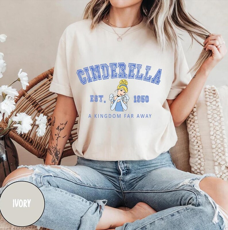 May include: A light beige t-shirt with a blue graphic of Cinderella. The graphic includes the text "Cinderella", "Est. 1950", and "A Kingdom Far Away".