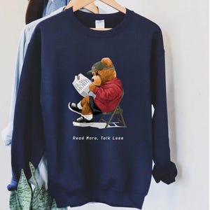 May include: A navy blue sweatshirt with a cartoon bear wearing a baseball cap and reading a newspaper. The bear is sitting on a red chair. The text "Read More, Talk Less" is printed below the bear.