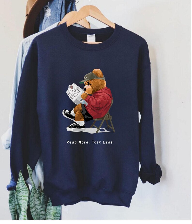 May include: A navy blue sweatshirt with a cartoon bear wearing a baseball cap and reading a newspaper. The bear is sitting on a red chair. The text "Read More, Talk Less" is printed below the bear.