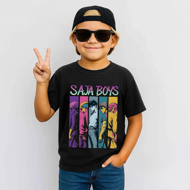 May include: Black t-shirt with the text "SAJA BOYS" in a colorful gradient font. Below the text is a graphic of five stylized figures in different colors. The person wearing the shirt is also wearing a black cap and sunglasses.