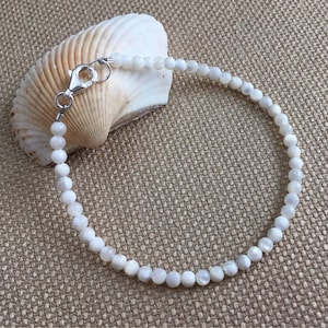 May include: A delicate white beaded bracelet with a silver clasp, resting on a white seashell. The bracelet is made of small, round, white beads. The clasp is silver and securely fastens the bracelet. The background is a textured, tan surface.